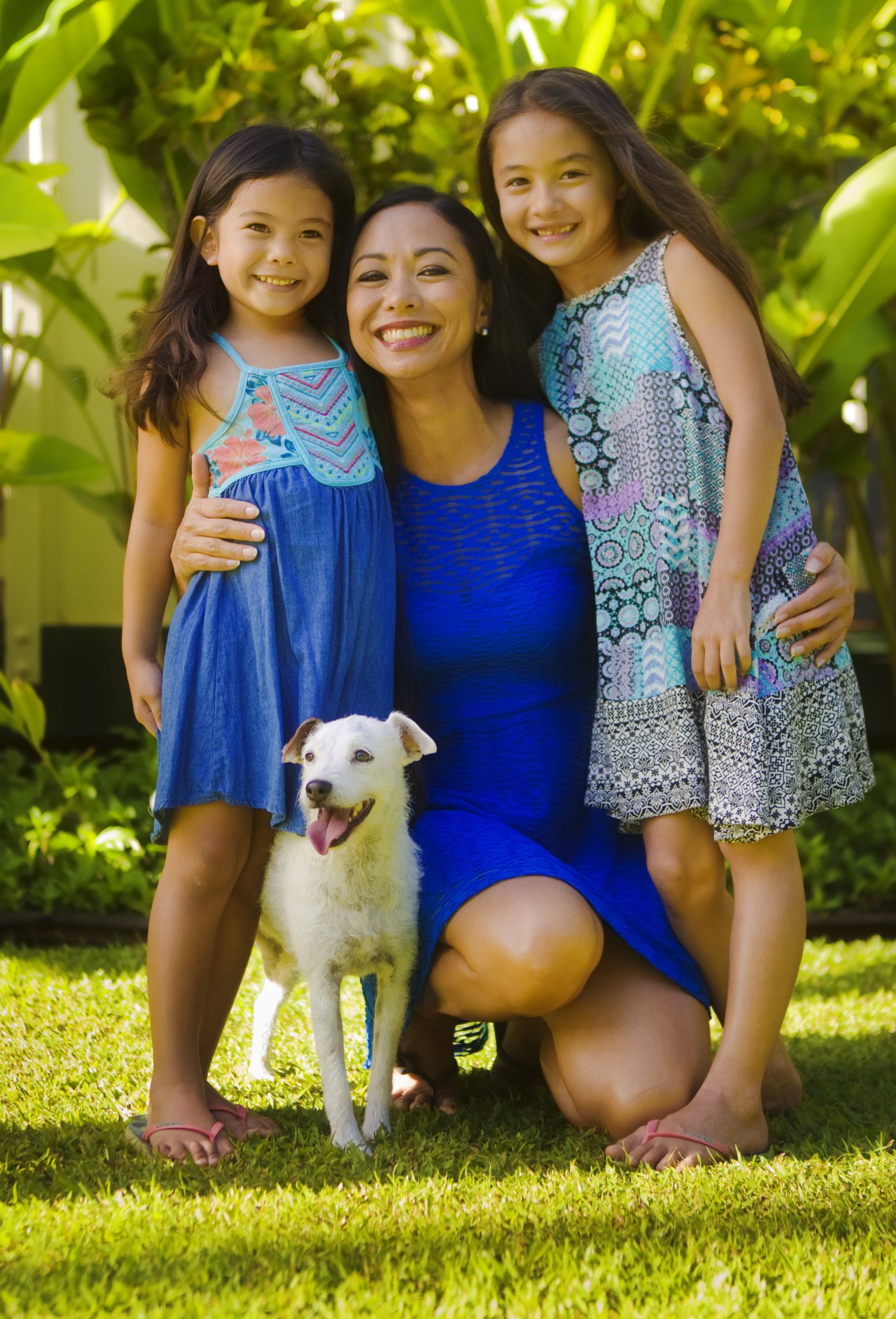 Portrait of a Hawaiian Polynesian family outdoor in their backyard. Two young girls with their mother and family pet dog, smiling and looking at camera full body group portrait.